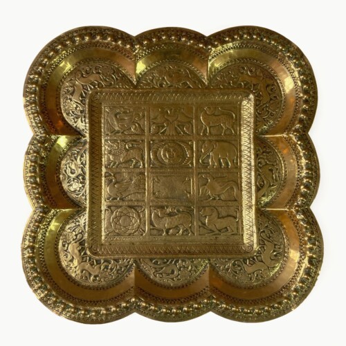An intricately designed metal tray on a white background. It is brass which is a dull golden colour and a bit shiny. The central part of the tray is a square with 12 rectangular sections in a grid with an ornate border. The outer part of the tray is 3 semi-circle shapes coming out of each side, making the silhouette have a cartoon cloud like shape. The Rectangular sections show symbols such as various animals, the sun and moon, and some more geometric and minimal ones.