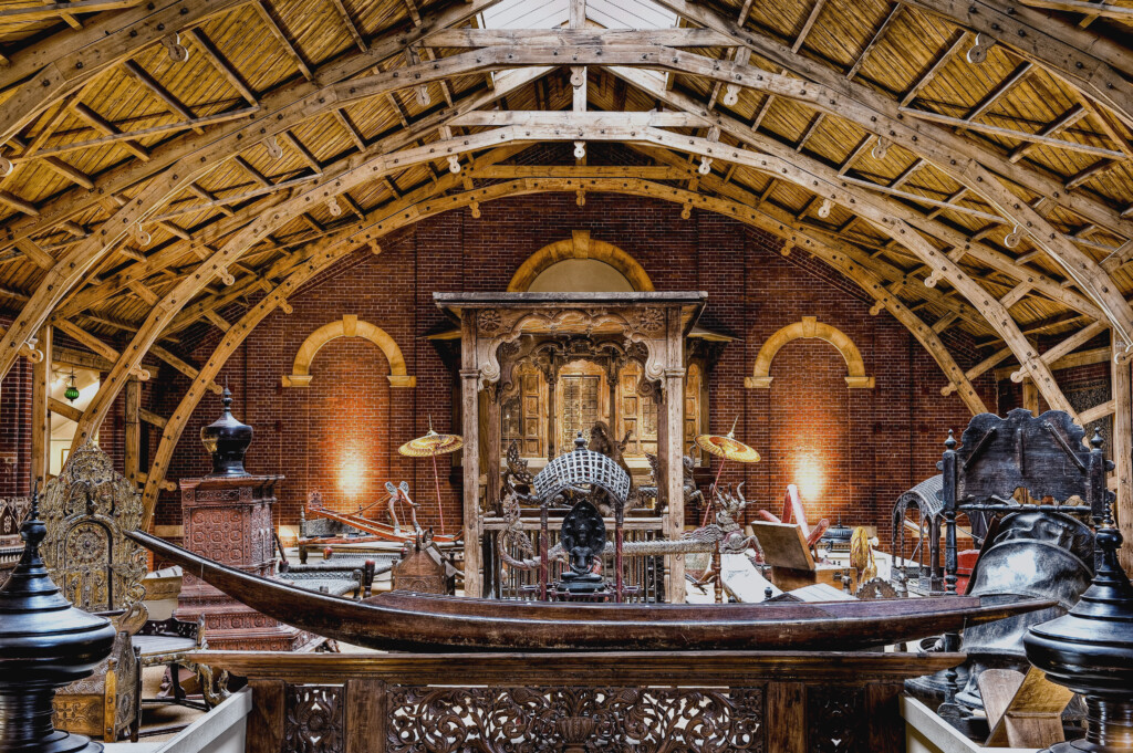 A photograph of the interior of The South Asia Collection. It shows the arched wooden roof, and a variety of mostly wooden ornate museum objects on a mezzanine in the middle.