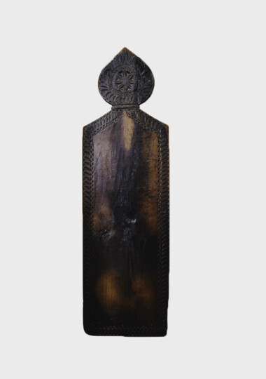 A vertical wooden prayer board. It is made of dark wood and you can see lighter patches where people have knelt and it is worn down. It is a rectangle with a rounded pointed part on the rop. There is some ornate carving in the top bit.