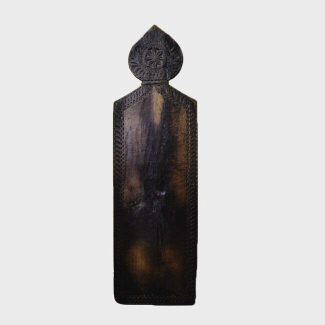 A vertical wooden prayer board. It is made of dark wood and you can see lighter patches where people have knelt and it is worn down. It is a rectangle with a rounded pointed part on the rop. There is some ornate carving in the top bit.