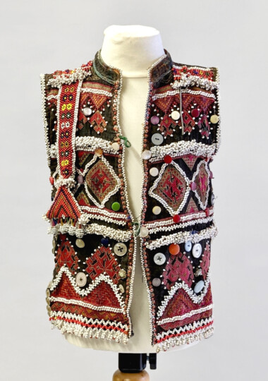 An intricate embellished child's waistcoat on a bust. It is red white and black in colour with lots of patterns, buttons, beadwork and stitching pattern throughout.