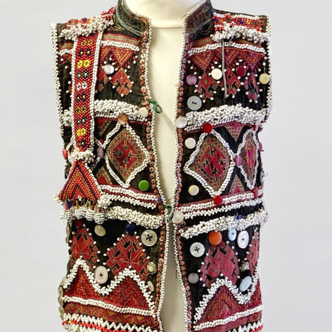 An intricate embellished child's waistcoat on a bust. It is red white and black in colour with lots of patterns, buttons, beadwork and stitching pattern throughout.