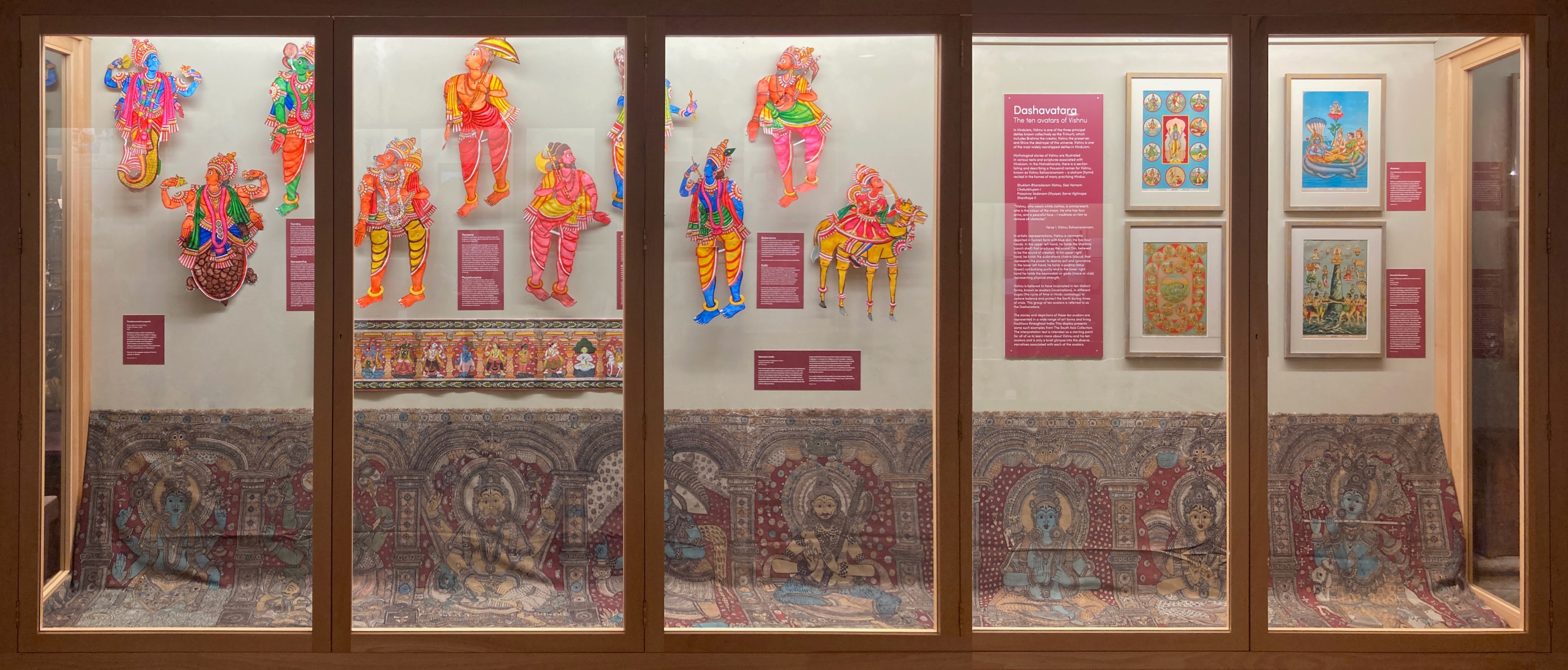 A photo of the full display case. It is a landscape case ,made up of five vertical windows with a light wood frame. The wall is painted an off white. There is a large wide textile taking the entire bottom third of the cabinet. This is a hand painted dark natural tone textile with intricate pattern and detail. It has eight deity figures on display. There is a similar acrylic painting that is a lot smaller on display above. There are ten brightly coloured puppets of the deity figures on display above on the left to middle of the cabinet. There are four colourful muted tone lithograph prints in a grid on the right. There is a large interpretation board in the middle of the puppets and lithographs and some smaller red labels around.