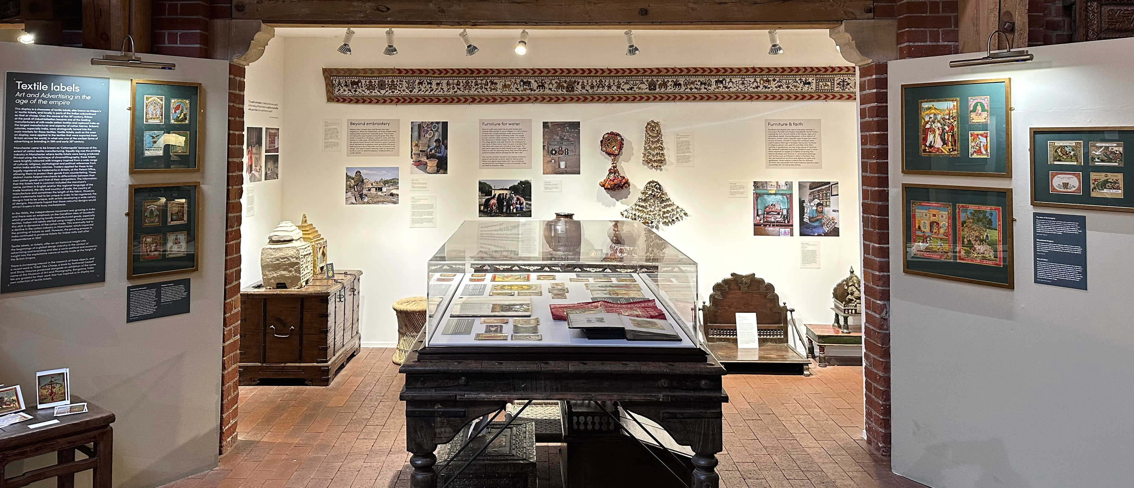 A view of the exhibition space showing a cloche in the centre two small green walls with pictures and text interpretation at either side and a different exhibition on a white wall with lost of different objects on display in the background.