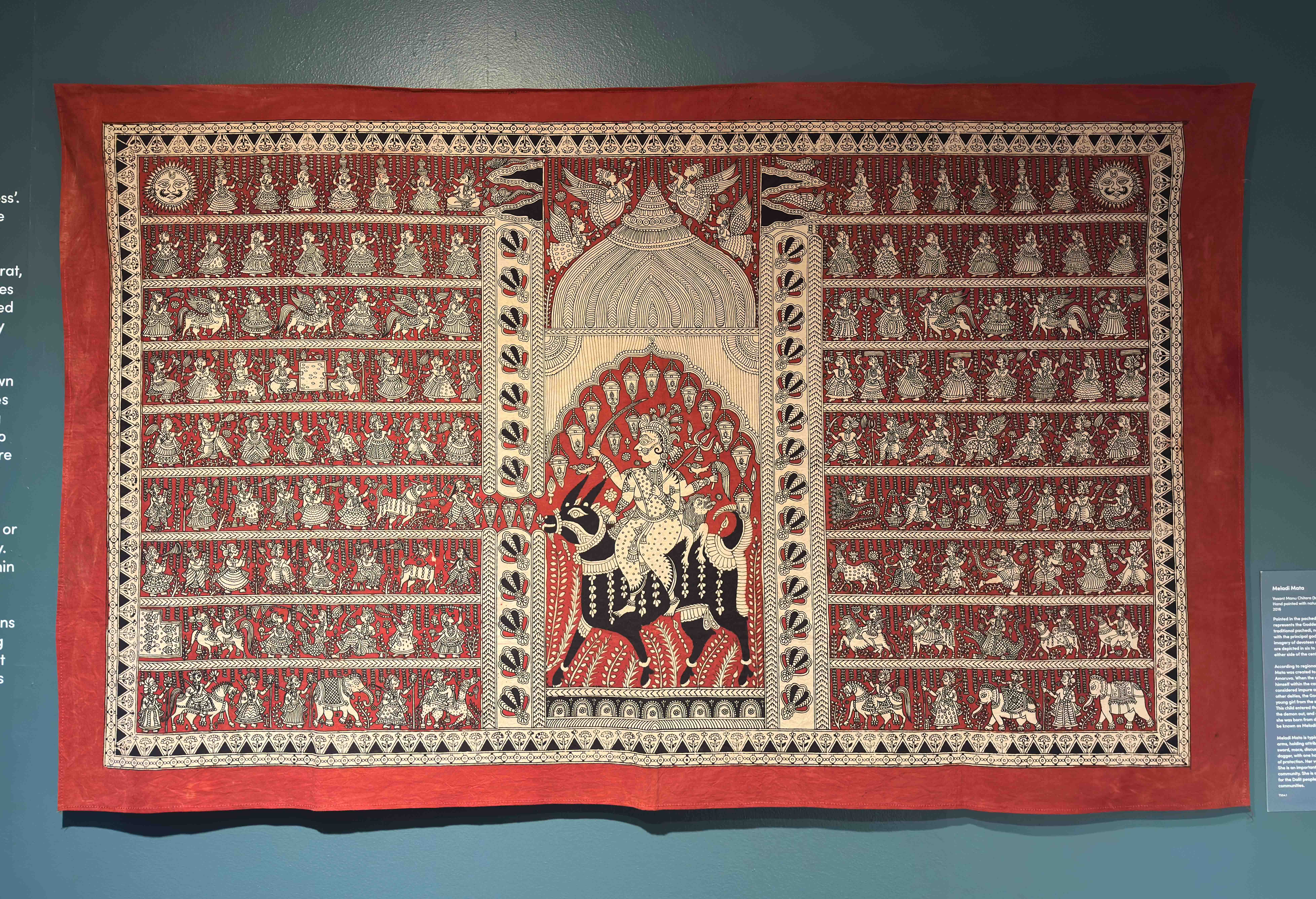 A view of a red, black and white textile on a blue wall. There is a goddess on a bull int the centre and lots of detailed figures around.