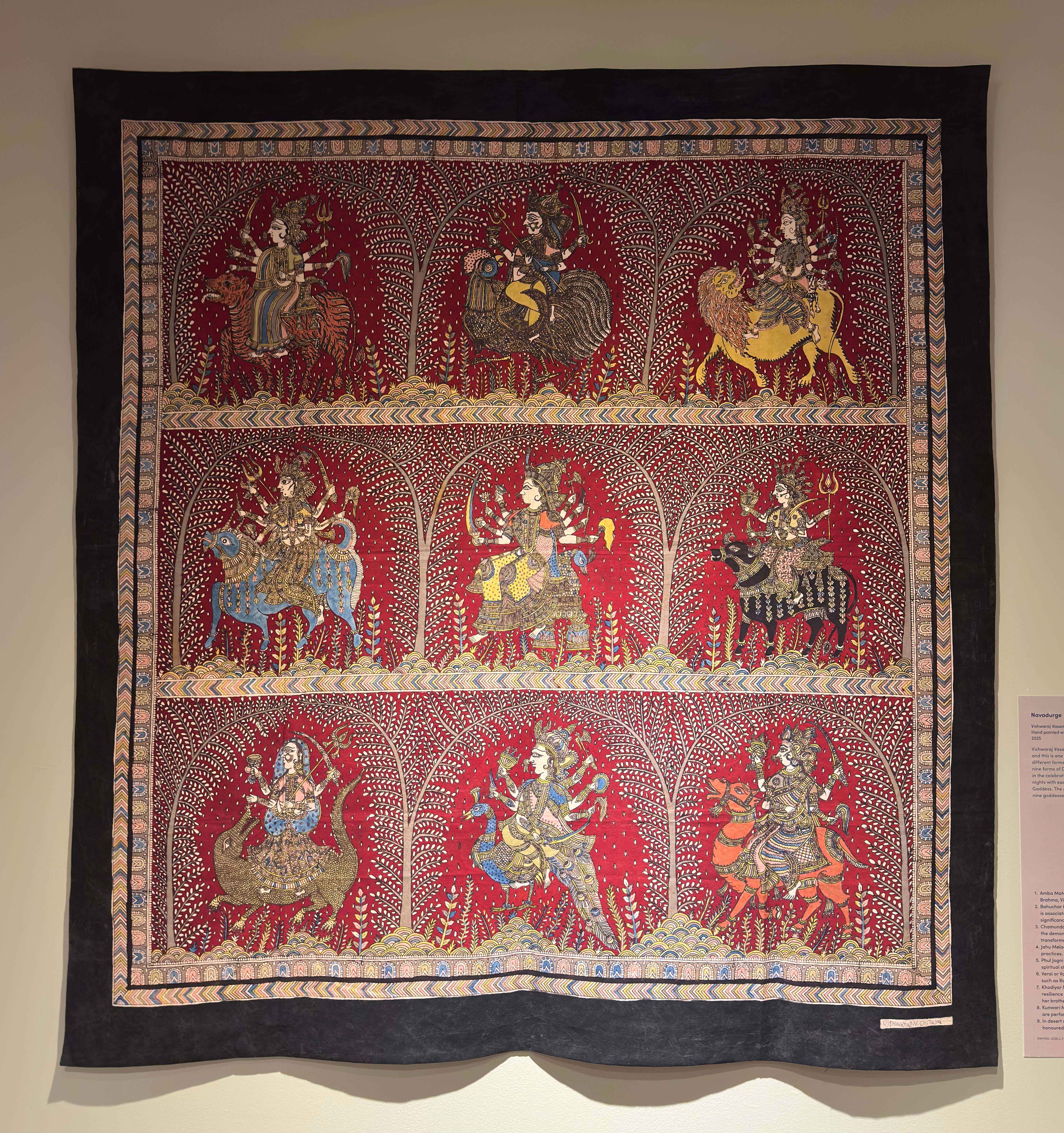 A textile titled Navadurge which displays a grid of 9 goddesses on a red background. Each with an animal mount and in fine detail.