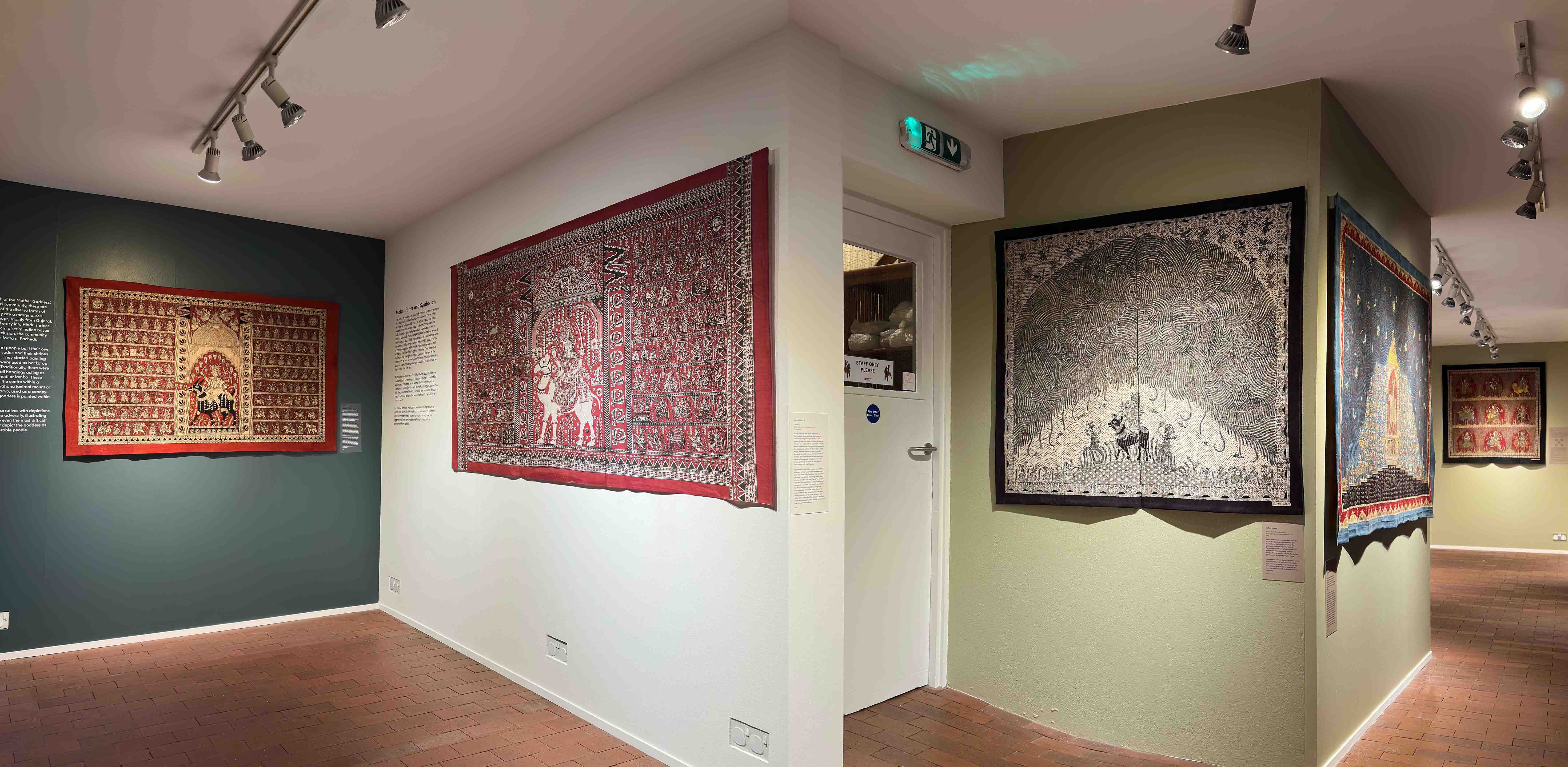 A view of the exhibition space showing several walls which are white, blue and green and each have very large detailed hand painted textiles on display.